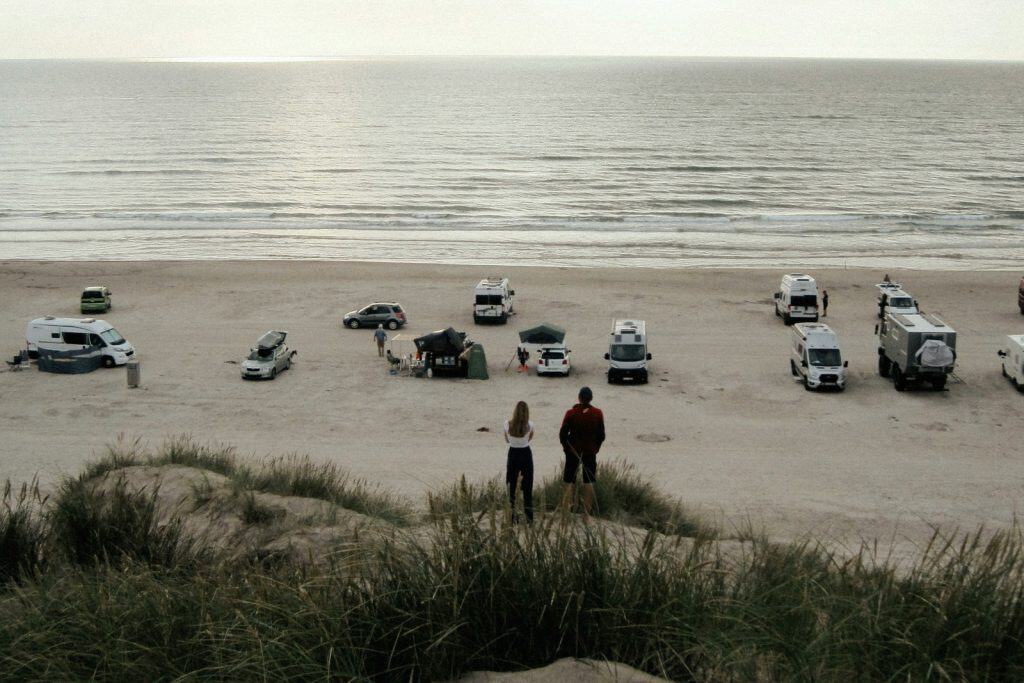 Zwei Personen beobachten den Strand mit mehreren geparkten Autos und Blick aufs Meer.