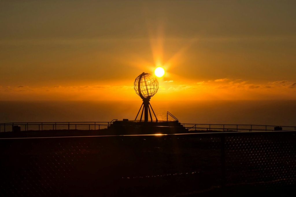 Sunset over water with a metal globe sculpture on a tripod on a platform, silhouetted against the orange sky.