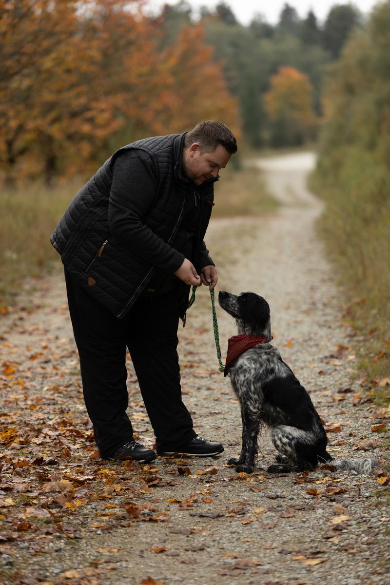 Mann in schwarzer Winterhose und Steppweste mit Hund auf einem Herbstweg, Seitenansicht mit Saum am Winterschuh