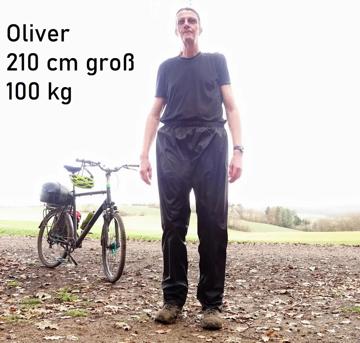 Oliver mit 210 cm Körpergröße trägt eine Regenhose mit 100 cm Innenbeinlänge neben seinem Fahrrad